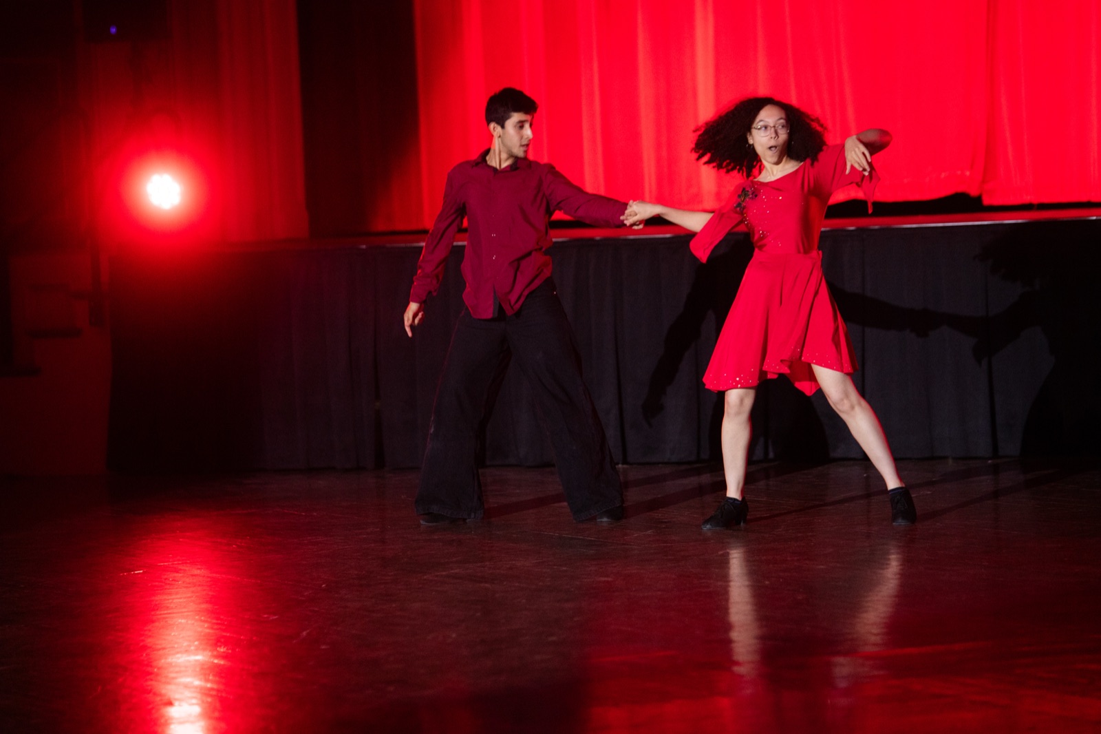 A couple in Latin open position on a red-lit stage, the follower in a red dress.